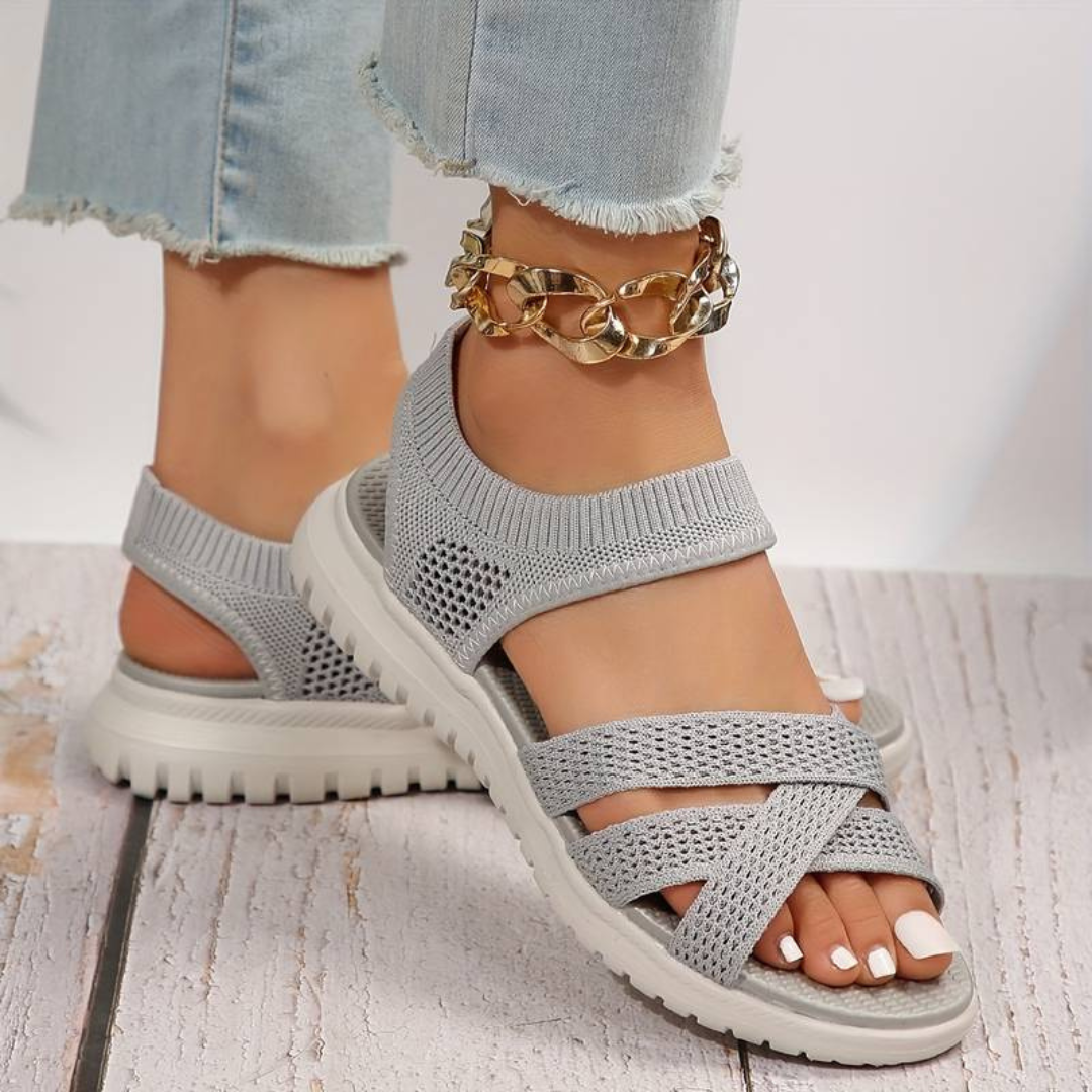 Graue Damen-Sandalen mit Strickdesign und dicker Sohle, getragen mit hellblauen Jeans und goldener Fußkette auf Holzboden.