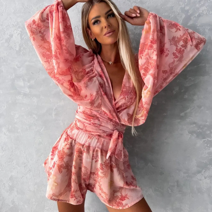 Frau in rosa geblümtem Jumpsuit vor grauer Wand, modisches Outfit, lange Ärmel, elegantes Design, trendige Damenmode, Sommerlook.