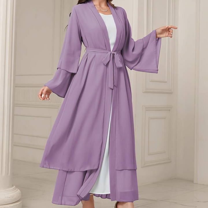 Frau in fliederfarbenem Kimono-Kleid mit Gürtel, stehend in elegantem Innenraum. Modetrend, Damenmode, stilvolle Kleidung, lila Kimono.