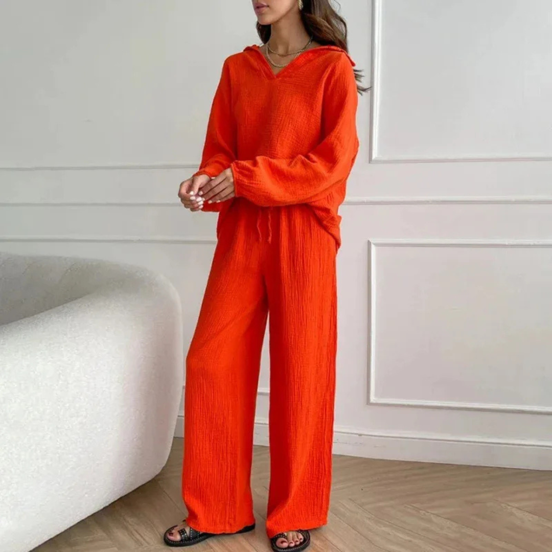 Frau in leuchtend orangefarbenem Freizeitanzug, bestehend aus Hoodie und Hose, steht in modernem Wohnzimmer. Modetrend, bequeme Damenbekleidung.