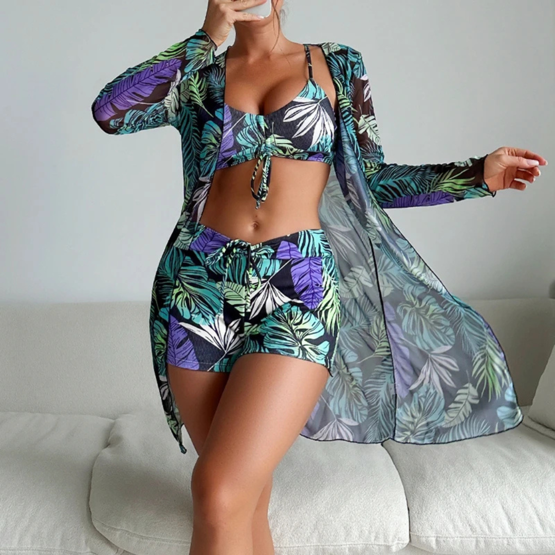 Frau in tropischem Badeanzug-Set mit Blättermuster, bestehend aus Bikini, Shorts und Kimono, posiert auf einem Sofa. Sommermode, Strandoutfit.