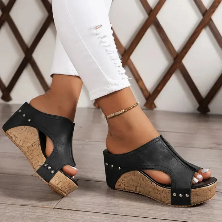 Schwarze Keilsandaletten mit Korksohle und Nieten, getragen von Person in weißen Jeans. Modische Sommerschuhe, bequem und stilvoll.