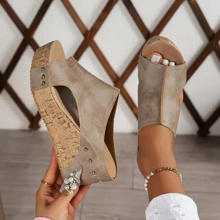 Beige Keilsandalen mit Korksohle und Nieten, getragen von einer Person mit Fußkettchen, vor einem Holzgitter-Hintergrund.