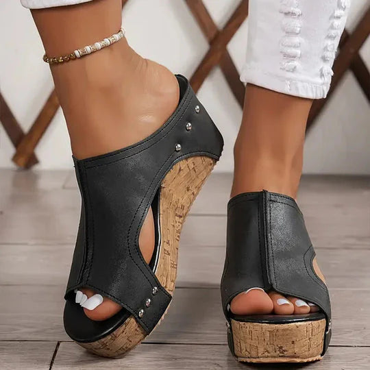 Schwarze Keilsandaletten mit Korksohle und Nieten, getragen von Frau in weißen Jeans. Modische Sommerschuhe, bequem und stilvoll.