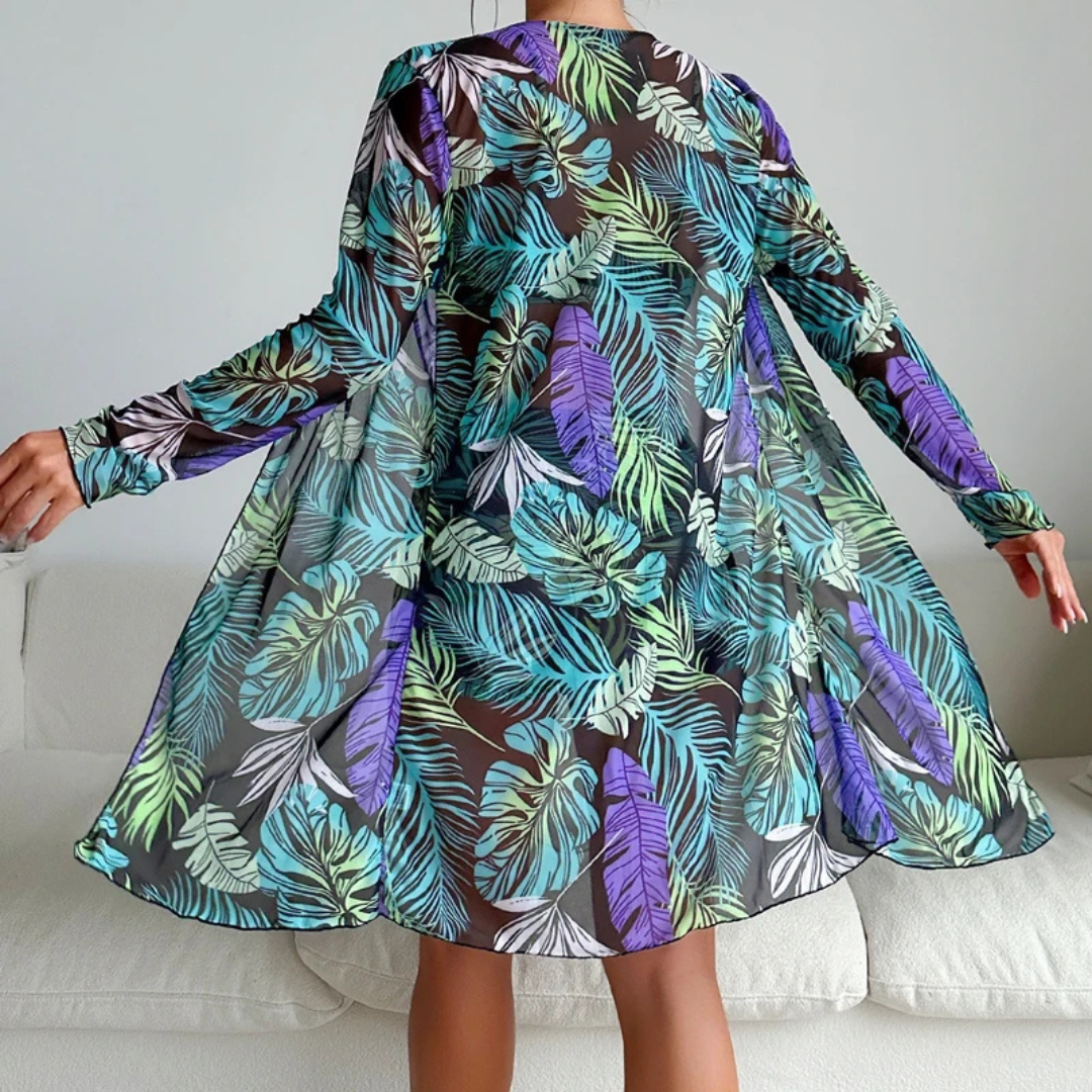 Frau in transparentem, langärmligem Kleid mit buntem Blätterdruck, stehend auf weißem Sofa. Modetrend, tropisches Design, Freizeitkleidung.