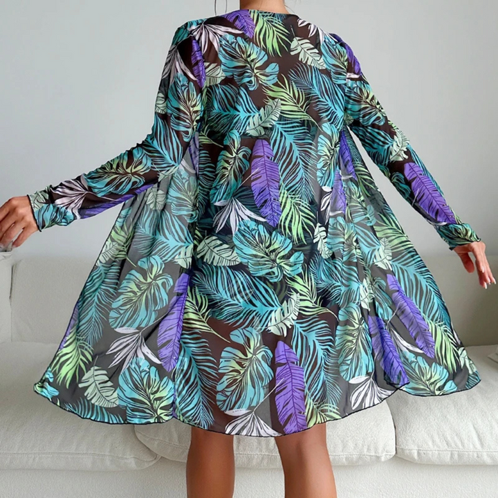 Frau in transparentem, langärmligem Kleid mit buntem Blätterdruck, stehend auf weißem Sofa. Modetrend, tropisches Design, Freizeitkleidung.