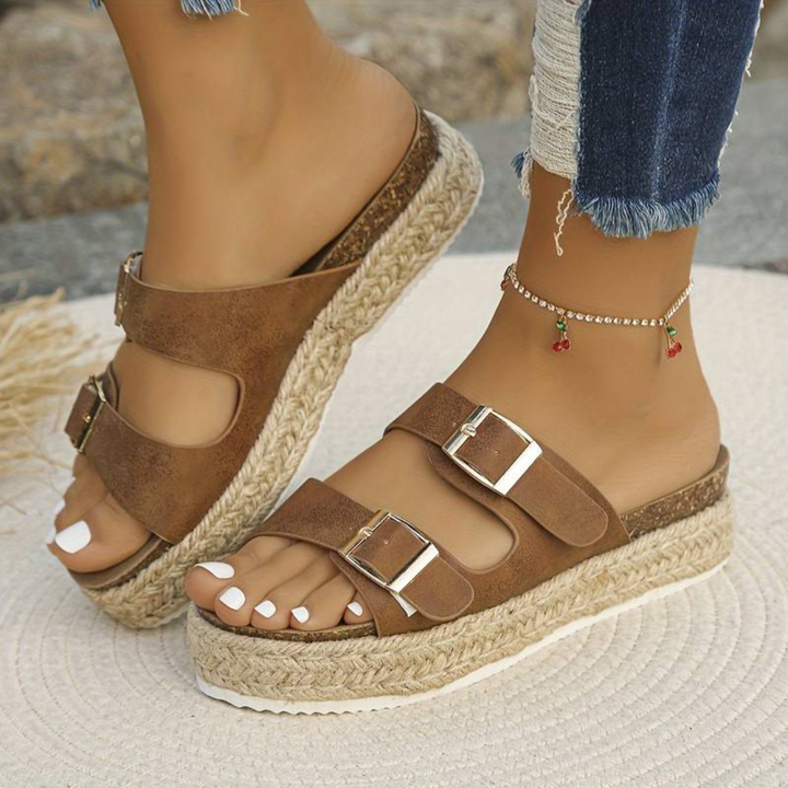 Braune Plateau-Sandalen mit Korksohle und Schnallen, getragen von einer Person mit Jeans und Fußkettchen. Sommermode, Damen-Schuhe, Freizeitlook.