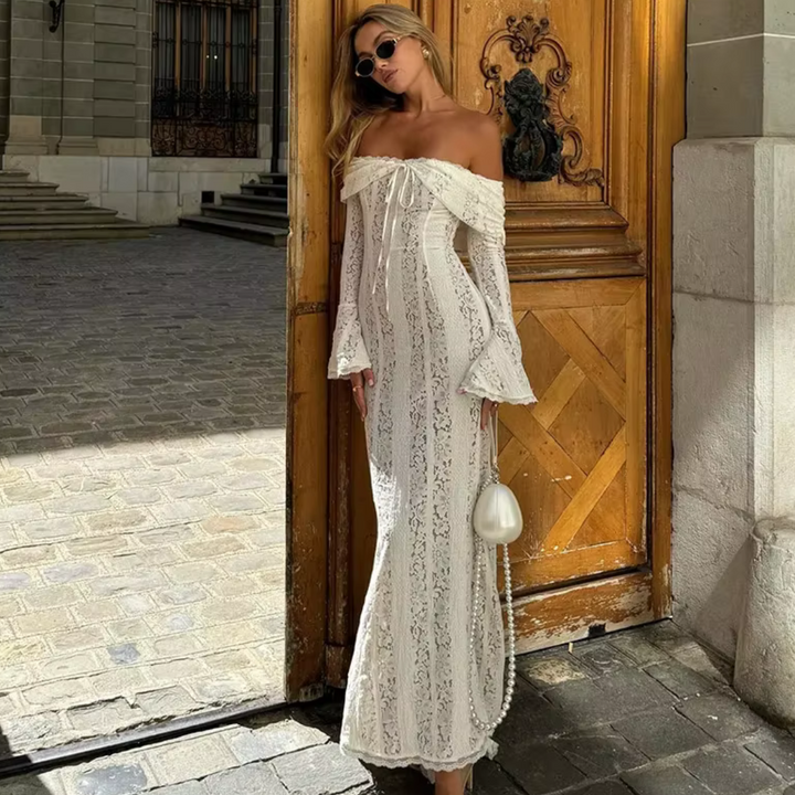Eine Frau in einem eleganten, schulterfreien weißen Spitzenkleid steht vor einer Holztür. Sie trägt Sonnenbrillen und hält eine kleine weiße Tasche.
