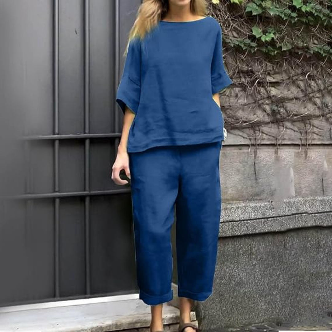 Frau in blauer Leinenkleidung, bestehend aus lockerem Oberteil und Hose, steht vor einer grauen Wand mit Efeu. Bequeme Sommermode, lässiger Stil.