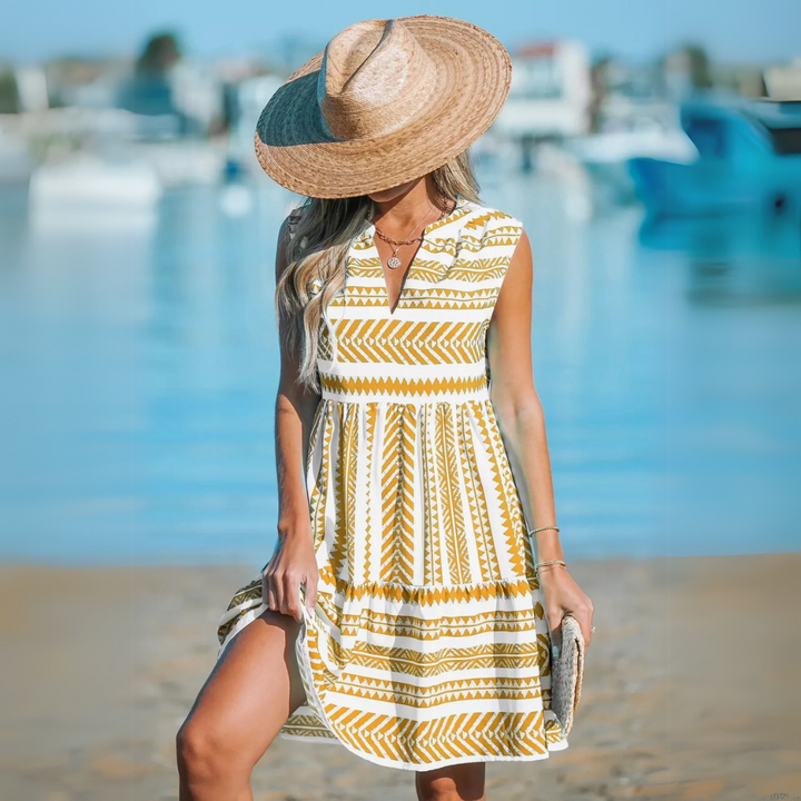 Frau in sommerlichem, gemustertem Kleid und Strohhut am Strand, mit verschwommenem Meereshintergrund. Perfekt für Sommermode und Strandoutfits.