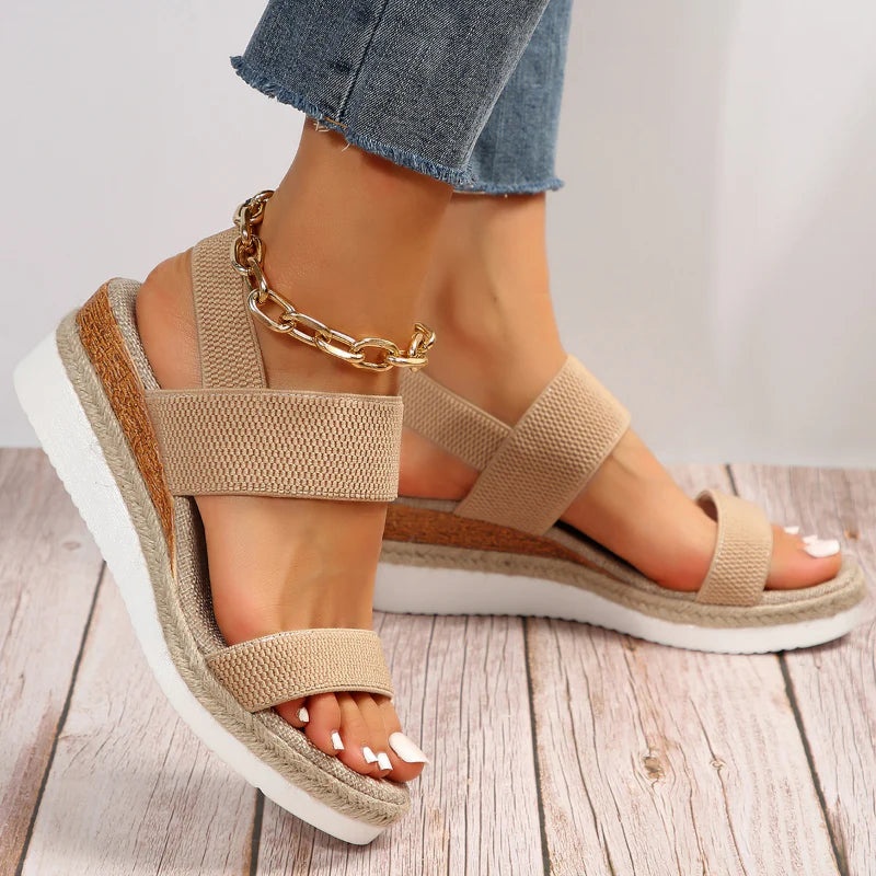 Beige Sandalen mit Keilabsatz und goldener Fußkette, getragen von einer Person in Jeans auf Holzboden. Modische Sommerschuhe für Damen.