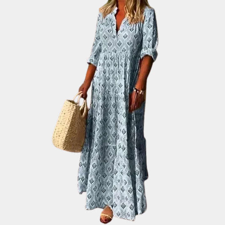 Langes, blau gemustertes Sommerkleid mit V-Ausschnitt und 3/4-Ärmeln, kombiniert mit Strohtasche und Sandalen. Ideal für Damenmode und Sommeroutfits.