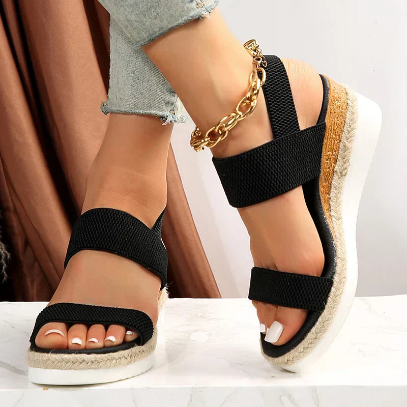 Schwarze Damen-Sandalen mit Keilabsatz, geflochtenem Juteband und goldener Knöchelkette, getragen mit hellblauen Jeans. Modische Sommerschuhe.