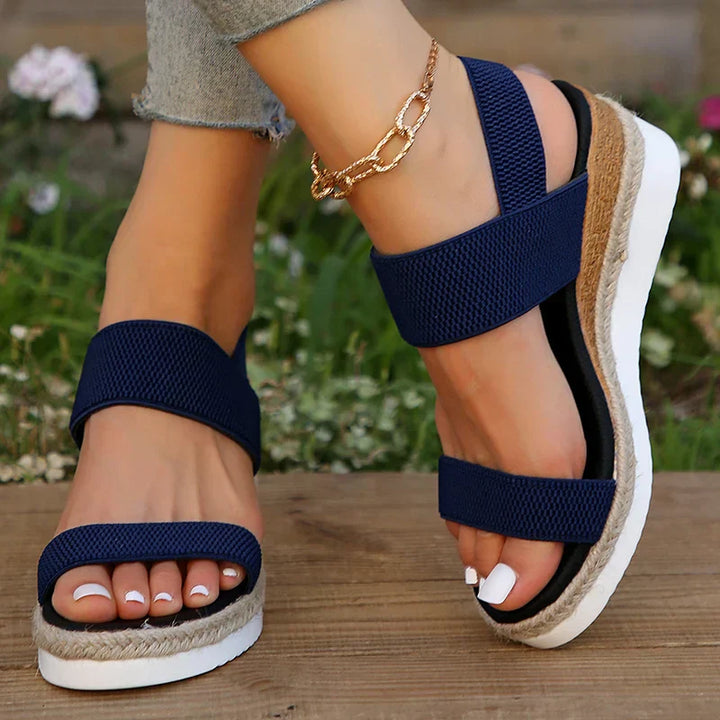 Damenfüße in blauen Sandalen mit Keilabsatz und goldener Fußkette auf Holzterrasse. Modische Sommerschuhe, bequem und stilvoll.