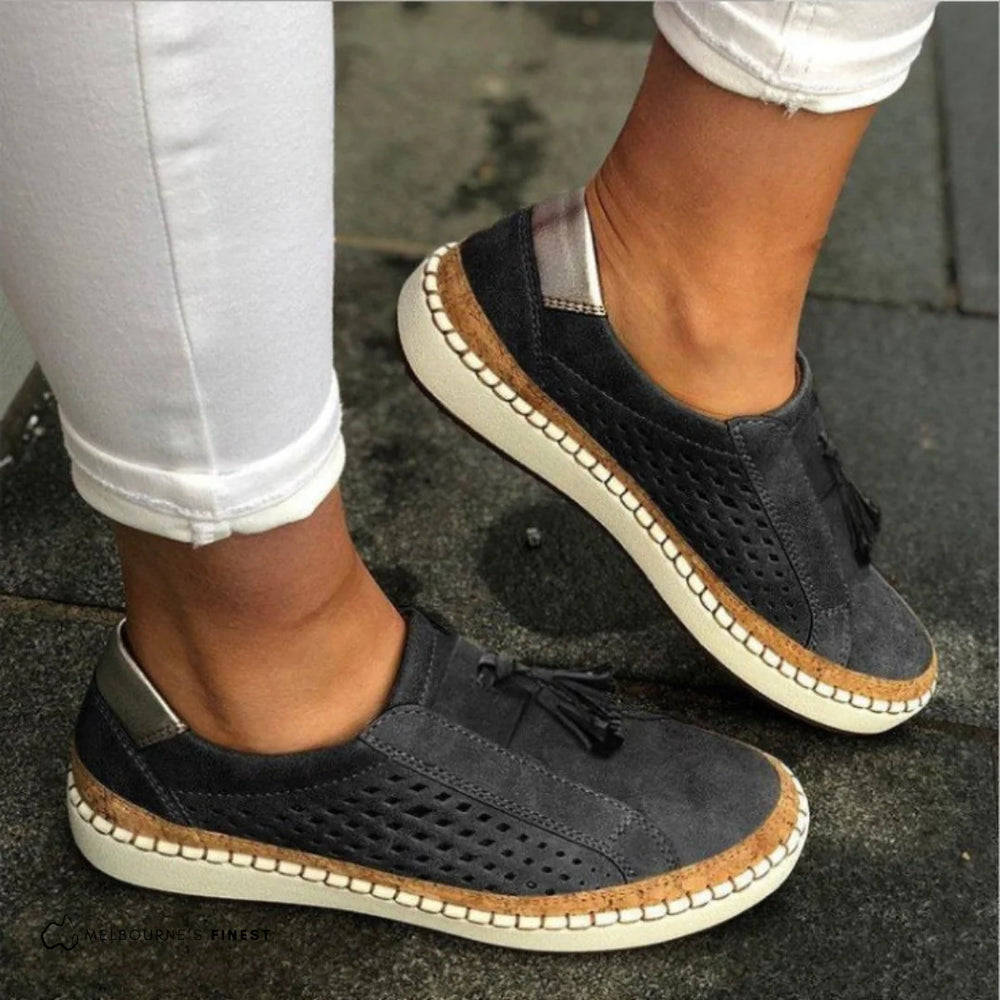 Schwarze Damen-Sneaker mit Lochmuster und Quasten, kombiniert mit weißen Hosen, auf grauem Pflaster. Bequeme Freizeitschuhe für den Alltag.