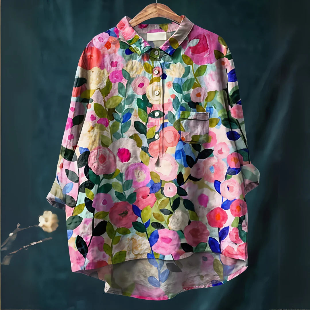Bunte Damenbluse mit floralem Muster, hängend auf einem Kleiderbügel. Lebendige Blumenmotive in Rosa, Blau und Grün auf dunklem Hintergrund.
