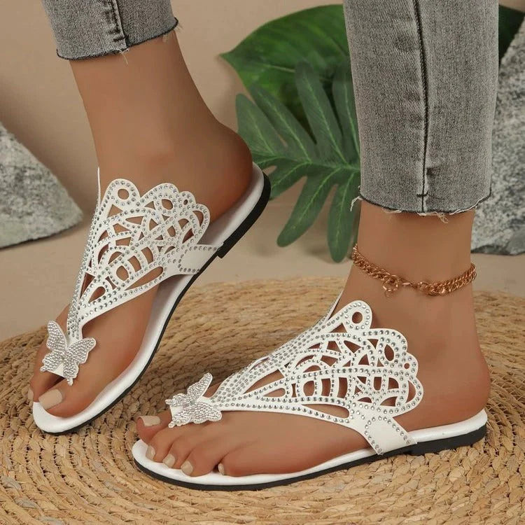 Weiße Sandalen mit Schmetterlingsdesign und Strasssteinen auf geflochtenem Teppich, getragen von Person in Jeans. Sommermode, Damen-Sandalen, elegant.