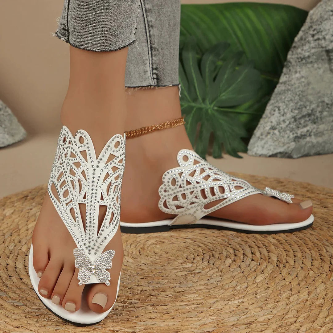 Damenfüße in weißen, verzierten Sandalen mit Schmetterlingsmotiv auf geflochtener Matte, graue Jeans, goldene Fußkette, tropische Blätter im Hintergrund.