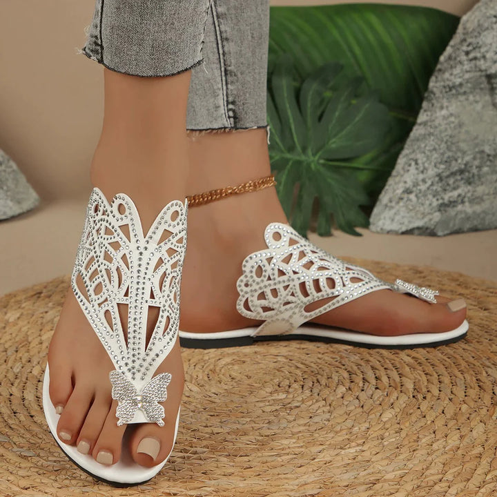 Damenfüße in weißen, verzierten Sandalen mit Schmetterlingsmotiv auf geflochtener Matte, graue Jeans, goldene Fußkette, tropische Blätter im Hintergrund.
