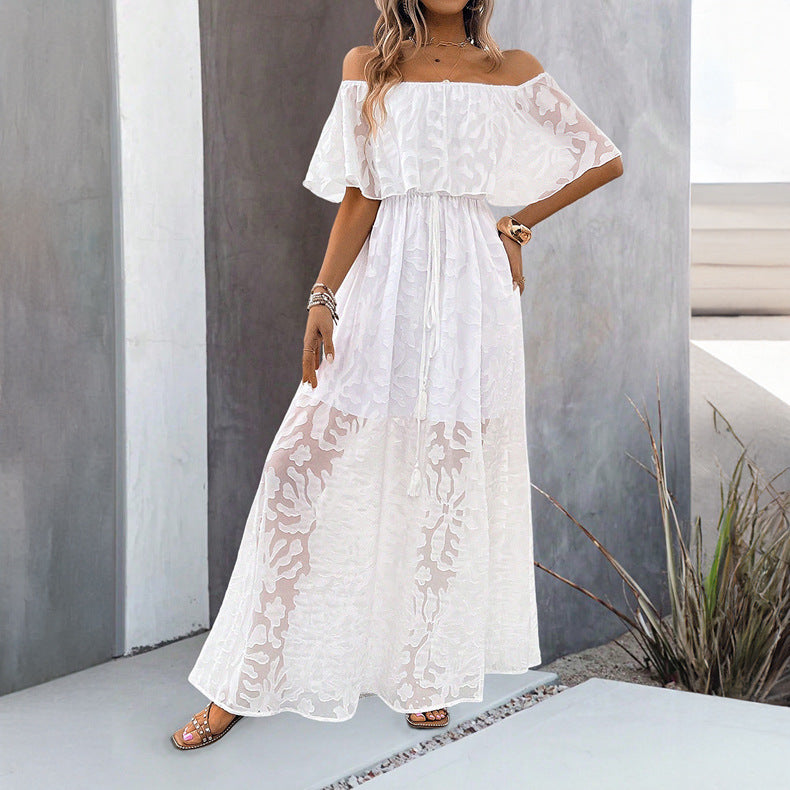 Frau in weißem, schulterfreiem Maxikleid mit Spitzenmuster, steht vor modernem Gebäude. Sommermode, elegantes Design, luftiger Stoff.