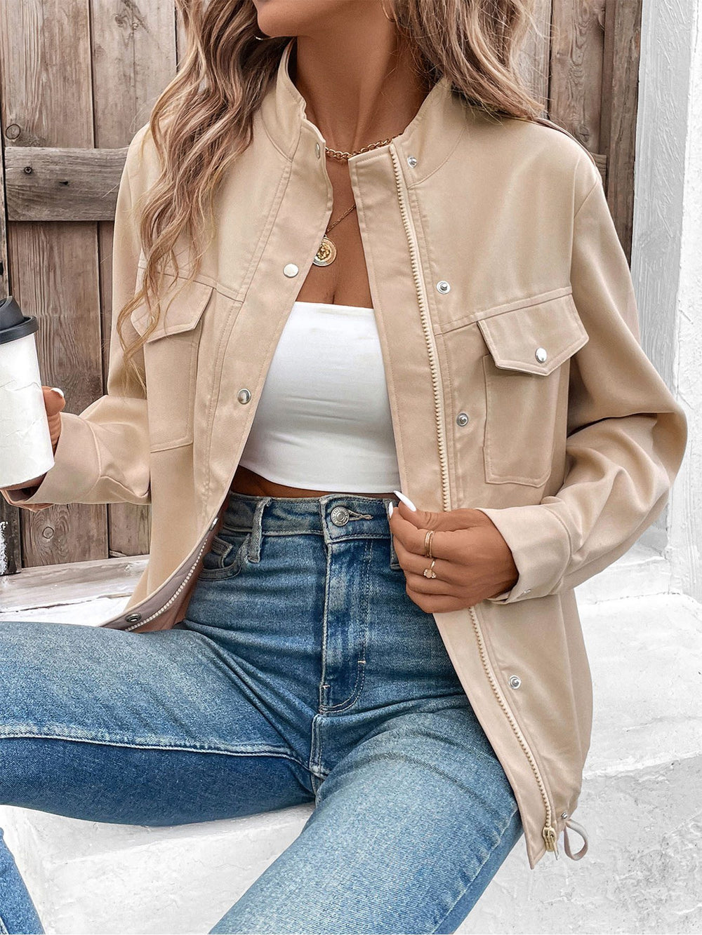 Frau in beiger Jacke und blauer Jeans, hält Kaffeebecher. Modetrend, lässiger Streetstyle, Herbstmode, Damenbekleidung, urbaner Look.