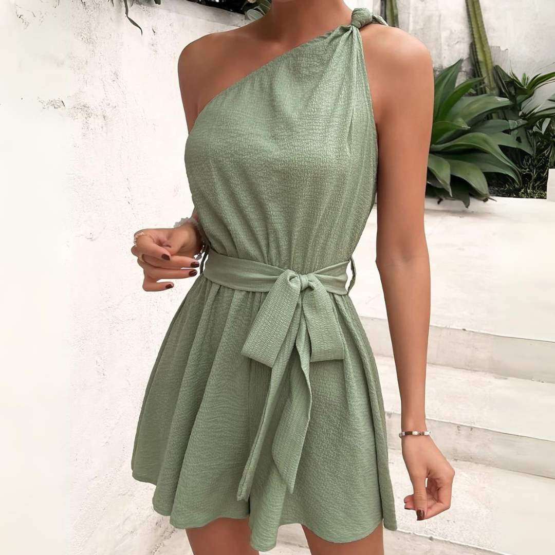 Frau in einem eleganten, einseitigen, grünen Sommerkleid mit Schleife, vor weißer Wand und Pflanzen. Modetrend, Damenmode, Sommeroutfit.