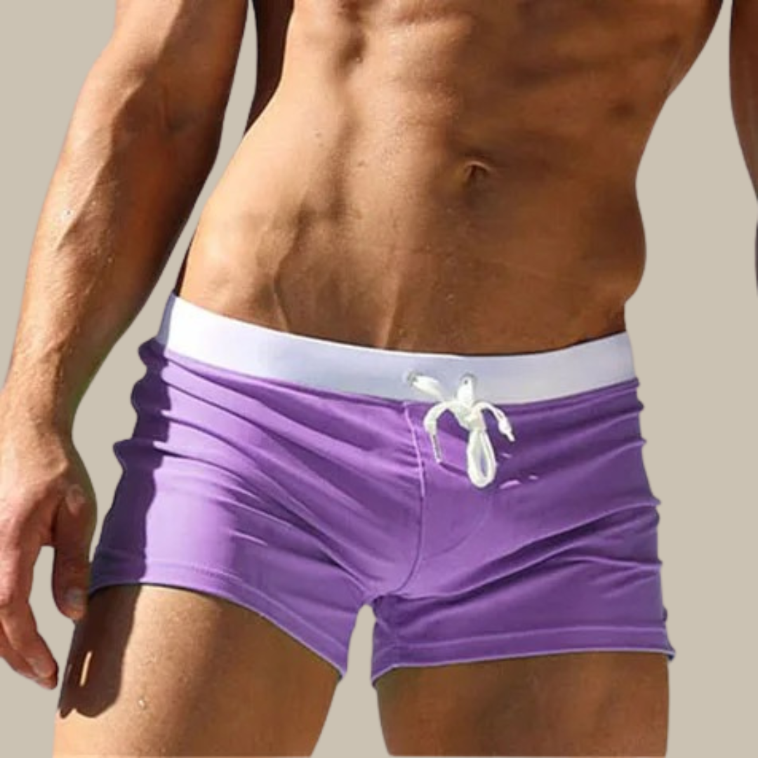 Mann in lila Badehose mit weißem Kordelzug, muskulöser Oberkörper, sonniger Hintergrund. Ideal für Strandmode, Sommer, Herrenbademode.