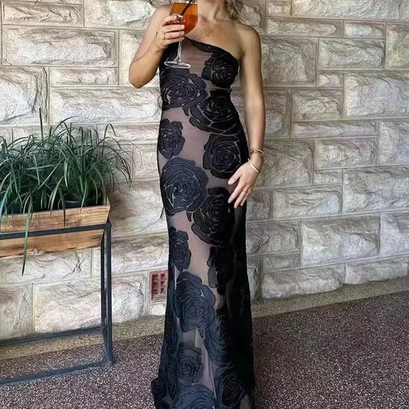 Frau in schwarzem, schulterfreiem Abendkleid mit Rosenmuster, hält ein Glas Wein. Elegante Mode vor Steinwand. Perfekt für Abendveranstaltungen.