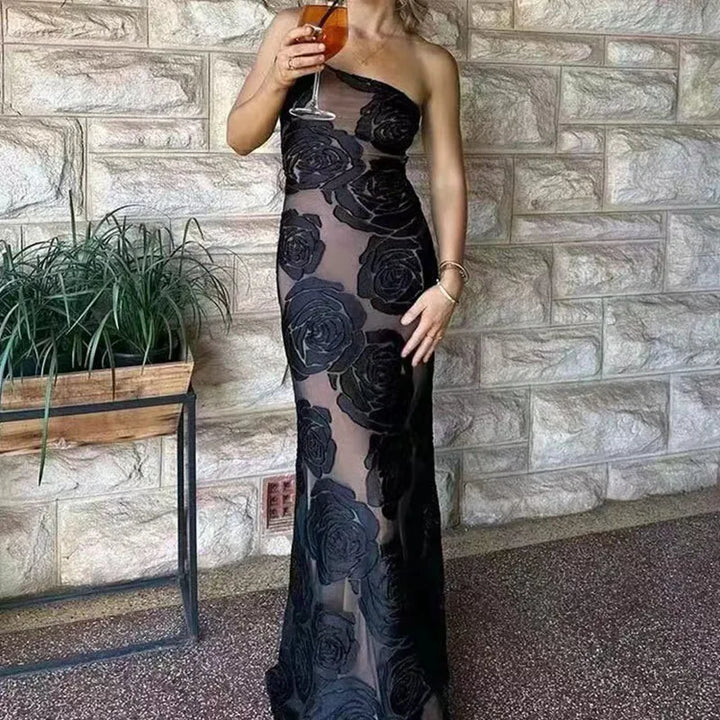 Frau in schwarzem, schulterfreiem Abendkleid mit Rosenmuster, hält ein Glas Wein. Elegante Mode vor Steinwand. Perfekt für Abendveranstaltungen.