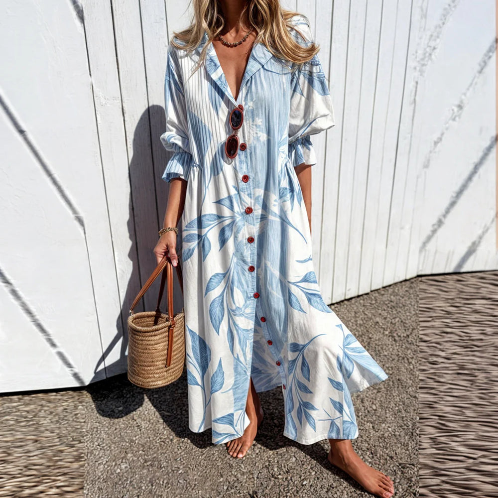 Frau in langem, geblümtem Sommerkleid mit blauen Blättern, trägt Strohtasche und Sonnenbrille, steht vor weißer Holzwand. Ideal für Strandmode.