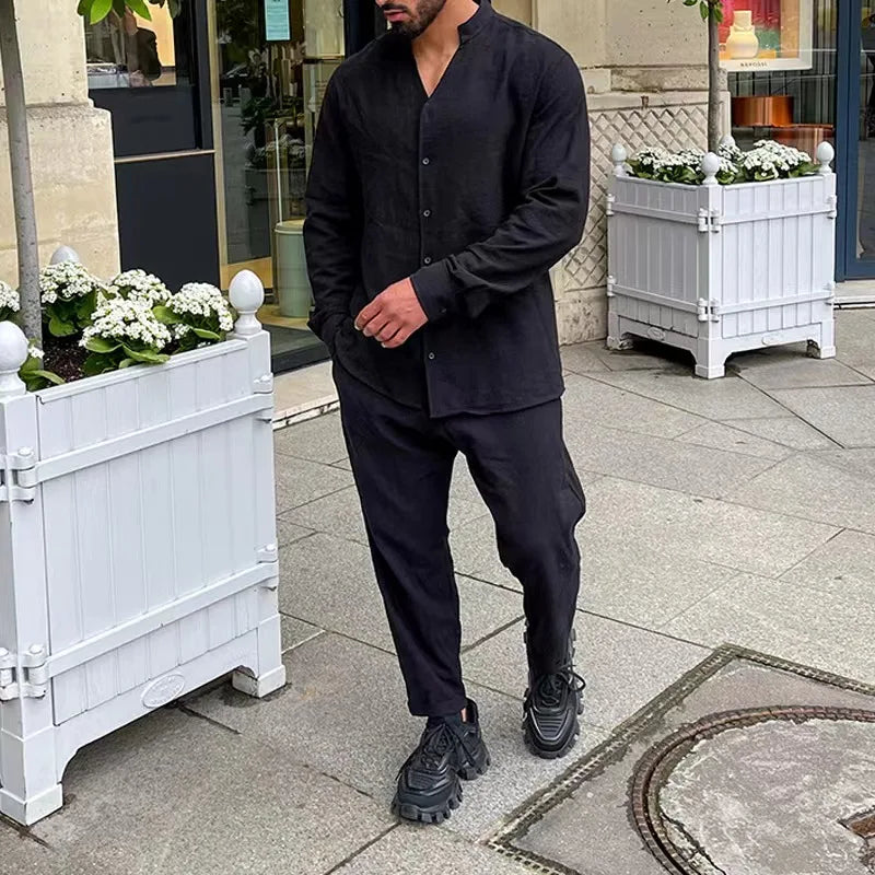 Mann in schwarzem Hemd und Hose, schwarze Sneaker, steht auf Gehweg vor weißem Pflanzkübel. Urbaner Stil, Herrenmode, lässiges Outfit.
