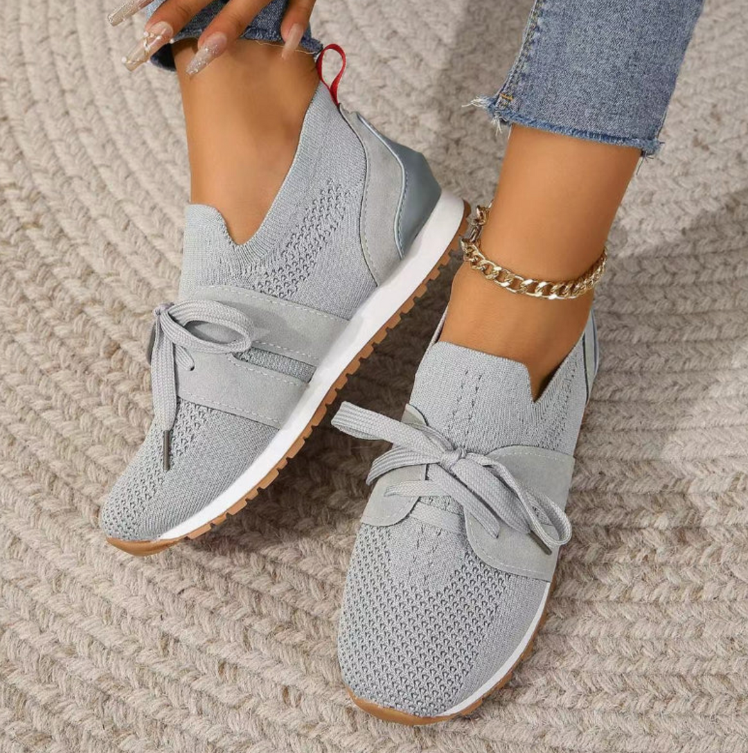 Graue Damen-Sneaker mit Strickmuster, weißen Sohlen und Schleifen, getragen mit blauen Jeans und goldener Knöchelkette auf geflochtenem Teppich.