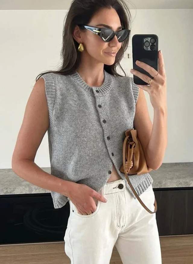 Frau in grauer Strickweste und weißer Hose macht Selfie mit Smartphone, trägt Sonnenbrille und goldene Ohrringe, hält braune Tasche.
