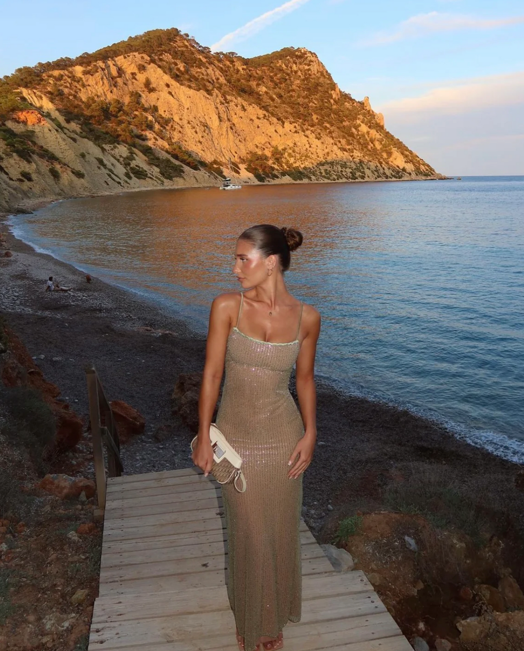 Frau in schimmerndem Abendkleid steht auf Holzsteg am Strand, mit malerischem Berg und Meer im Hintergrund bei Sonnenuntergang.