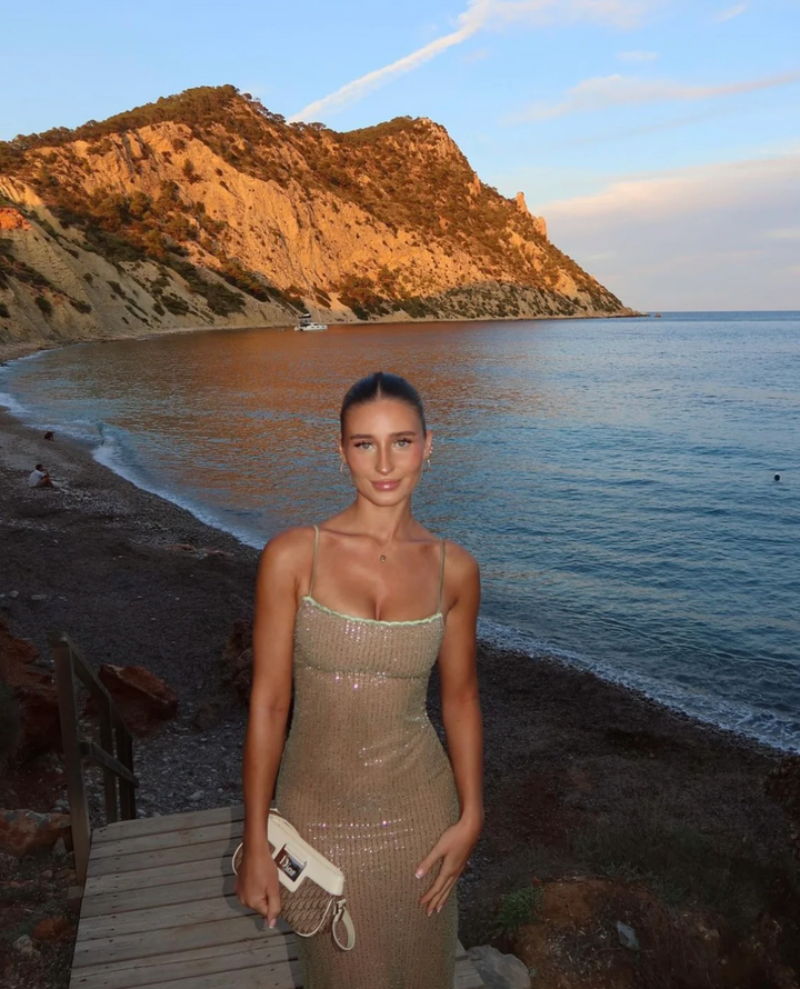 Frau in glitzerndem Kleid am Strand von Ibiza bei Sonnenuntergang, mit felsiger Küste im Hintergrund. Sommermode, Strandurlaub, Ibiza-Küste.