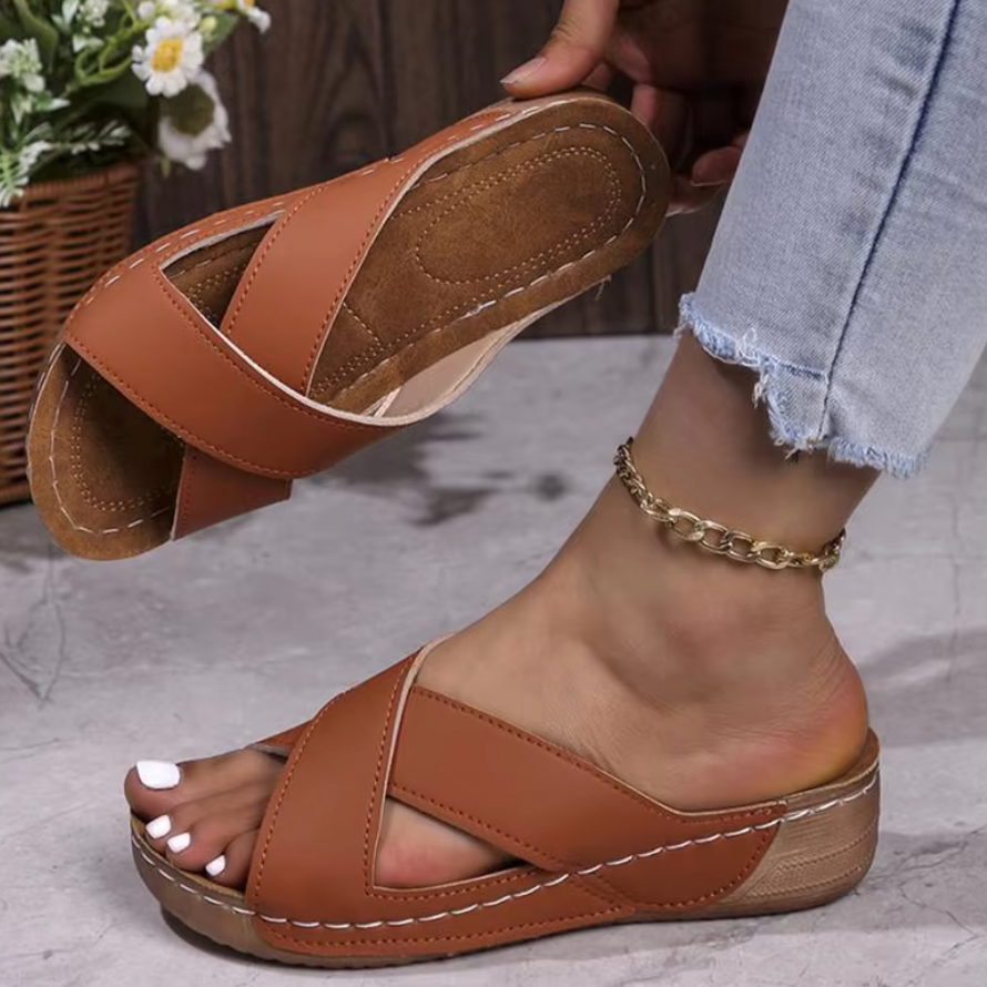 Braune Damen-Sandalen mit Kreuzriemen, getragen mit Jeans und goldener Fußkette. Bequeme Sommerschuhe auf grauem Boden, Blumen im Hintergrund.