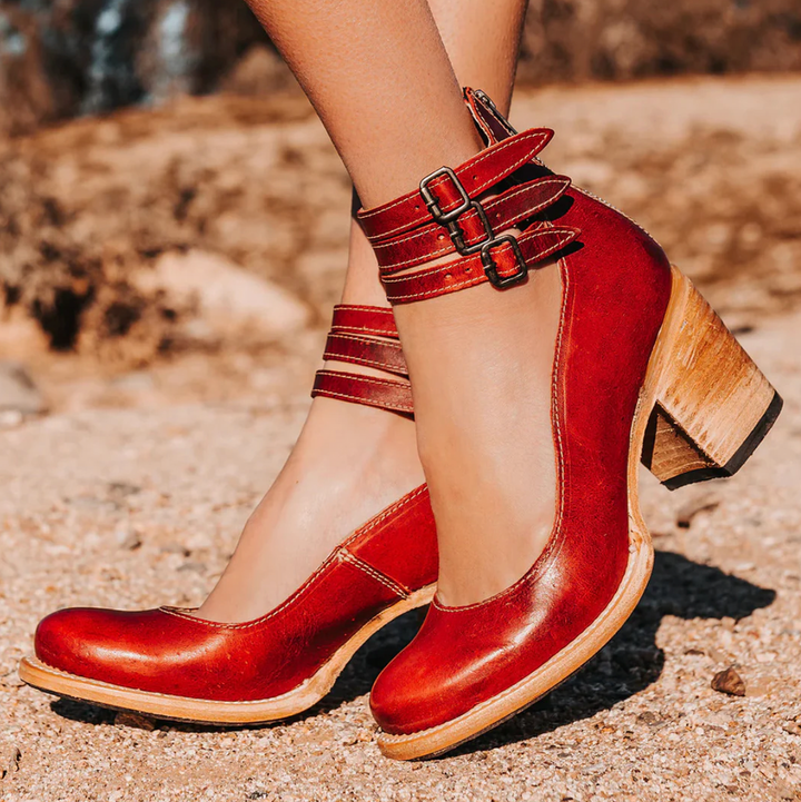 Rote Damen-Lederpumps mit Blockabsatz und Riemen, stilvoll auf sandigem Untergrund präsentiert. Elegante Schuhe für modische Anlässe.
