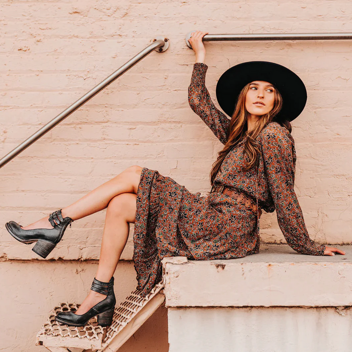 Frau in geblümtem Kleid und schwarzem Hut sitzt lässig auf Treppe. Modetrend, Herbstmode, Damenbekleidung, Streetstyle, Boho-Chic.