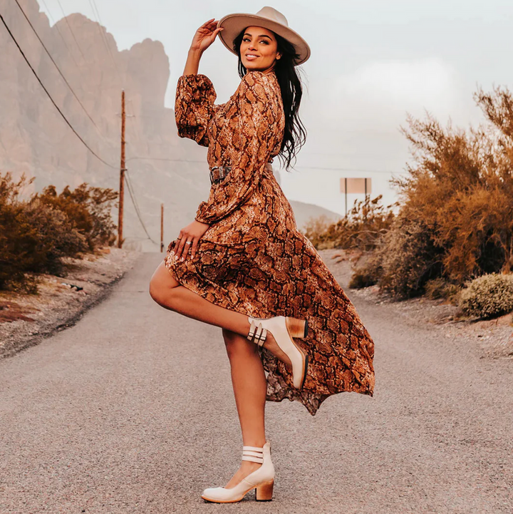Frau in langem Schlangenmuster-Kleid und Hut posiert auf ländlicher Straße. Modetrend, Boho-Stil, Herbstmode, Outdoor-Fotoshooting.
