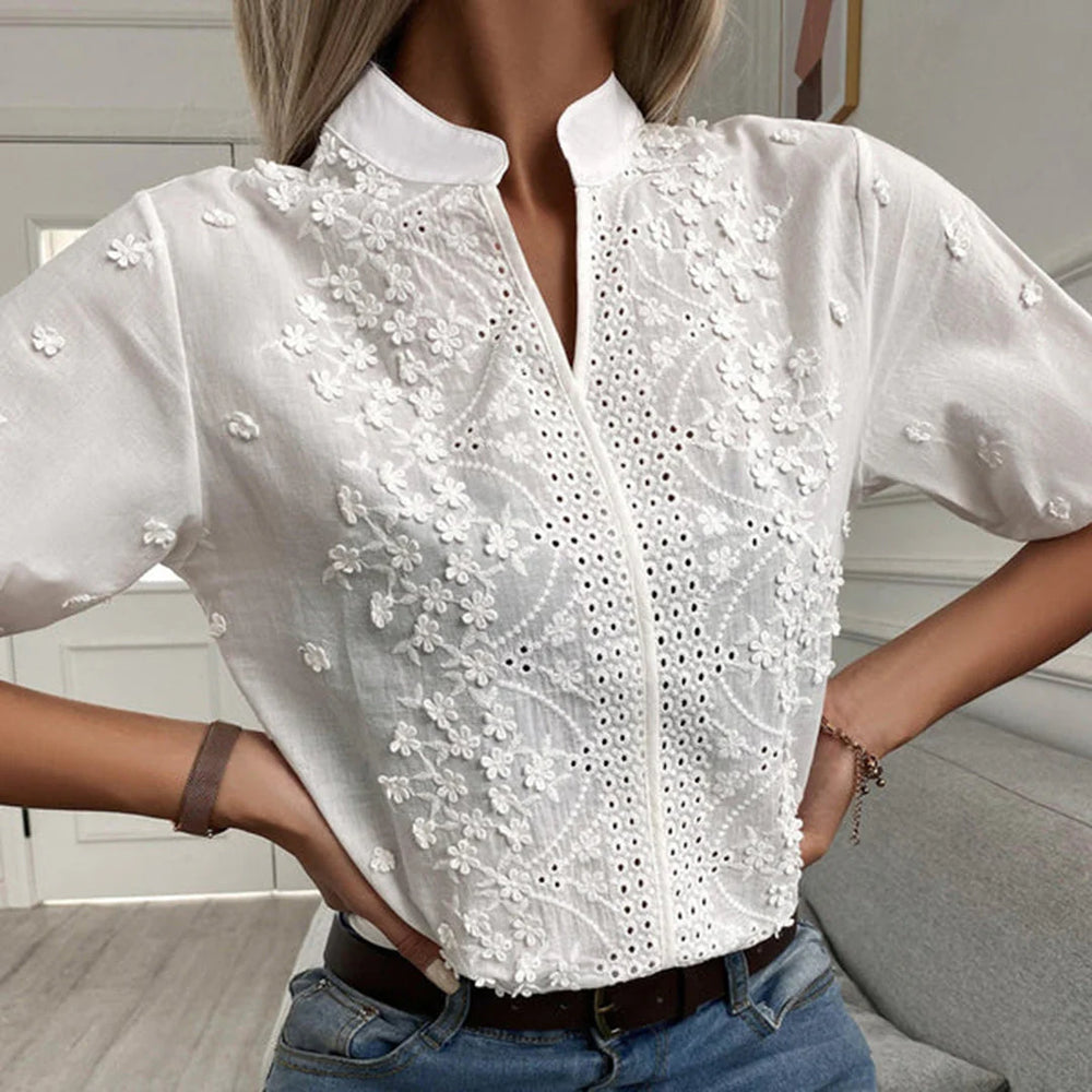 Frau trägt weiße, bestickte Bluse mit Blumenmuster, Stehkragen und kurzen Ärmeln. Modetrend, elegante Damenbekleidung, Sommeroutfit.