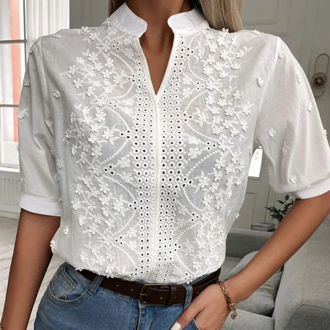 Frau trägt weiße, bestickte Bluse mit floralen Mustern und Lochstickerei, kombiniert mit blauer Jeans. Elegante Damenmode, Sommeroutfit.