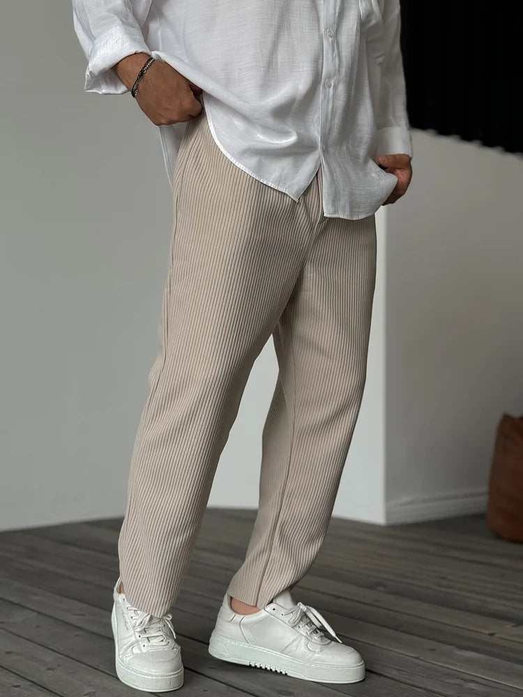 Mann in beiger Plissee-Hose und weißem Hemd, trägt weiße Sneaker. Moderne Herrenmode, lässiger Stil, elegante Freizeitkleidung.