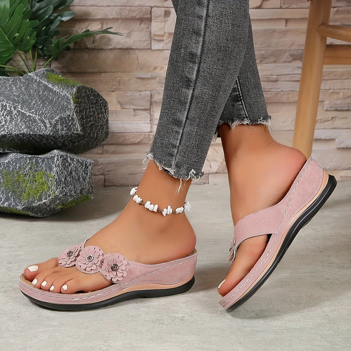 Damenfüße in rosa Sandalen mit Blumenmuster, graue Jeans, Fußkettchen, Steinwand im Hintergrund. Bequeme Sommerschuhe, modische Damenmode.