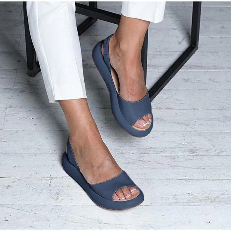 Blaue Damen-Sandalen mit offenem Zeh, getragen von einer Person in weißen Hosen, auf hellem Holzboden. Bequeme Sommerschuhe für Frauen.