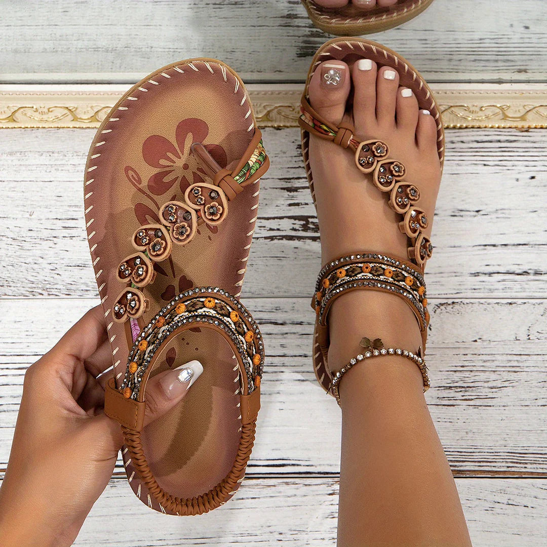 Braune Sandalen mit Blumenmuster und Perlenverzierung auf Holzuntergrund, Damenhand hält einen Schuh. Sommermode, bequeme Fußbekleidung.