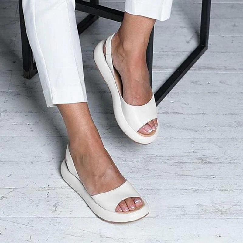Weiße Damen-Sommerschuhe mit offenem Zeh, getragen von einer Person in weißen Hosen, auf hellem Holzboden. Bequeme, stilvolle Sandalen.