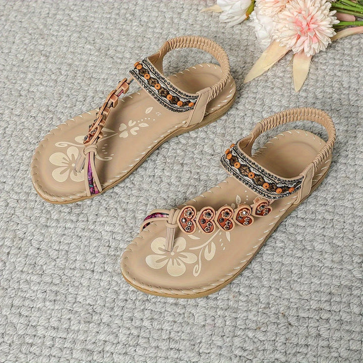 Beige Sandalen mit Blumenmuster und bunten Perlen auf grauem Teppich, dekoriert mit rosa Blumen. Ideal für Sommermode und Freizeitkleidung.