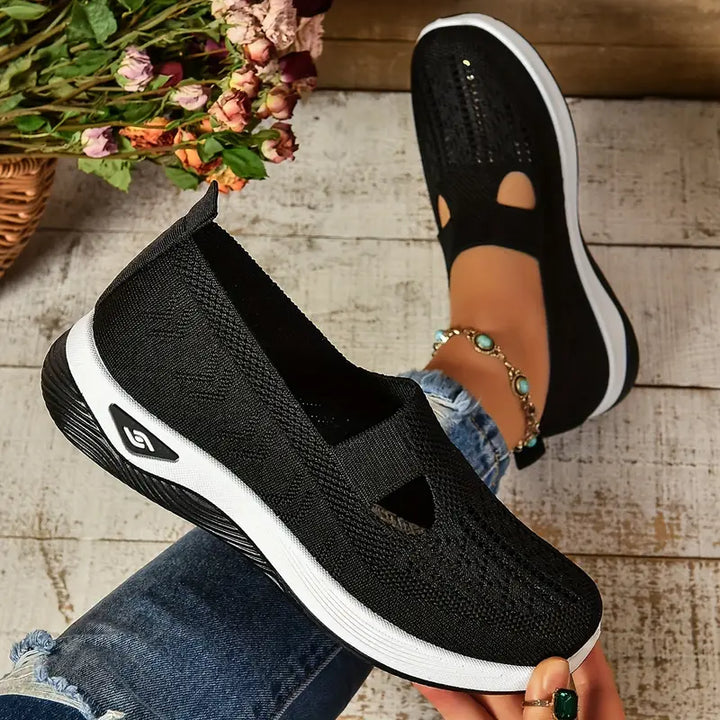 Schwarze Damen-Sneaker mit weißer Sohle, atmungsaktivem Strickmaterial, getragen von Person mit Jeans und Blumenarmband, auf Holzboden.