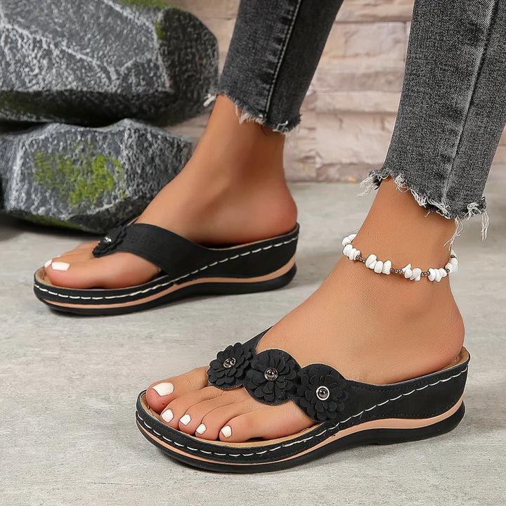Schwarze Damen-Sandalen mit Blumenverzierung, getragen mit grauen Jeans und weißem Fußkettchen. Bequeme Sommerschuhe für Damen.