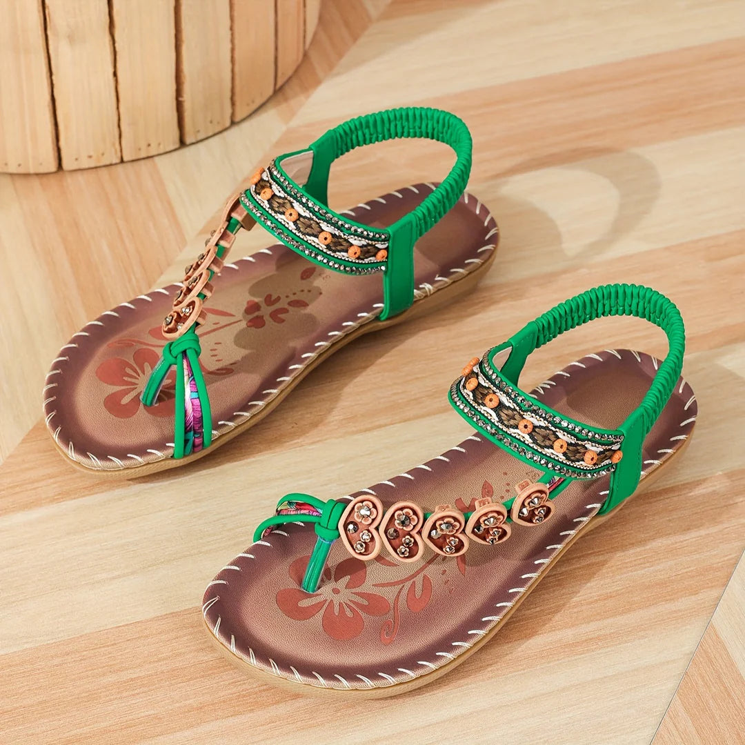 Grüne Damen-Sandalen mit Blumenmuster, verziert mit Perlen und Strasssteinen, auf Holzfußboden. Sommermode, bequeme Riemchensandalen.